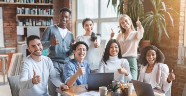 Neşeli, çoklu etnik çeşitlilikte genç bir iş ekibi ofiste birlikte poz veriyor ve başparmaklarını kaldırıyor.