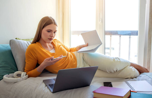A teen girl relaxes on her bed, balancing her laptop and notes while focused on her studies. Sunlight streams through the window, enhancing the serene atmosphere.