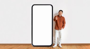 Asyalı adam kocaman boş bir akıllı telefonun yanında duruyor, gülümsüyor ve başparmağını kaldırıyor, ahşap döşemeli parlak ve minimalist bir ortamda heyecan ya da onayı işaret ediyor..