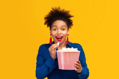 Free Time. Portrait of excited black girl eating popcorn and looking at camera, watching movie isolated over yellow wall