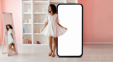 Şık beyaz bir elbise giyen genç siyah bir kadın, şık, yumuşak renkli bir odada, görüntüsünü yansıtan, büyük, boş bir telefon ekranının yanında sevinçle duruyor..