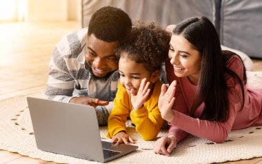 Ev izolasyonu sırasında çevrimiçi iletişim. Anne ve kızı ellerini dizüstü bilgisayarda sallıyor, baba gülümsüyor.