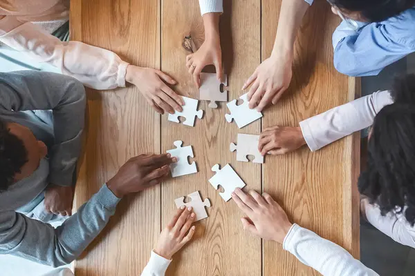 Collaboration concept, top view of creative multiracial team sitting around table and putting together puzzle pieces, cropped