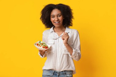 Sağlıklı beslenme. Gülümseyen afro kadın sebze salatası yiyor, sarı arka planda duruyor fotokopi alanı ile ve kameraya bakıyor.
