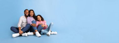 Gülümseyen mutlu Afro-Amerikan ebeveynler genç ergen bir kızla yerde oturuyor, mavi arka planda izole edilmiş kızına sarılıyorlar, panoramik çekim, bedava fotokopi alanı, afiş.