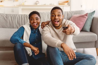 Heyecanlı Afrikalı siyahi çift TV şovu ya da film izliyor, ellerinde uzaktan konsolla evde yerde oturuyor ve oturma odasındaki kanepeye yaslanıyor, kameraya bakan şaşkın adam ve kadın.