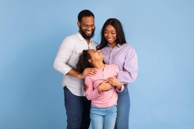 Aile Aşk Konsepti. Mavi stüdyo arka planında izole edilmiş, gülümseyen kıvırcık kıza sarılan Afrikalı erkek ve kadının portresi. Anne ve baba kameraya kızlarıyla poz veriyorlar.