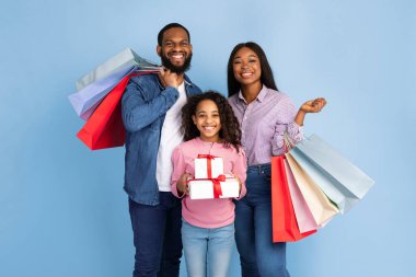 Tatil Hediyeleri Alışveriş. Parlak alışveriş torbaları tutan üç kişilik neşeli siyah aile portresi. Heyecanlı genç kız, paketlenmiş hediye kutuları tutuyor. Turkuaz stüdyo duvarında izole edilmiş.