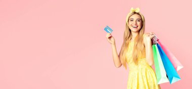 İnternetten alışveriş. Heyecanlı sarışın kız elinde kredi kartı ve kese kağıtlarıyla pembe stüdyo duvarında, kopyasal alanda