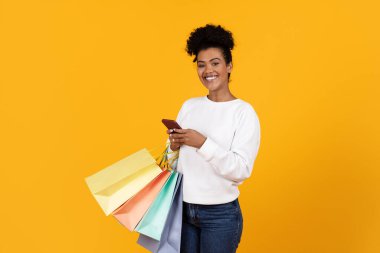 Neşeli genç bir kadın, bir elinde renkli alışveriş çantaları, diğer elinde telefonuyla canlı sarı bir arka planda duruyor ve alışveriş gününün tadını çıkarıyor..