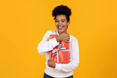 Kıvırcık saçlı neşeli bir kadın gümüş kurdeleye sarılmış parlak kırmızı bir hediyeyi kucaklıyor. Canlı sarı bir arka planın önünde duruyor, heyecan ve mutluluk saçıyor..