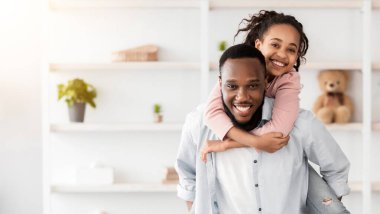 Baba ve kızı. Sevgi dolu genç bir siyah adam, mutlu bir kızı sırtında taşırken ve kameraya gülümserken, tatlı bir çocuk, babalarının omuzlarında gezerken, evde birlikte eğlenirken, kameraya bakarken,