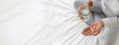 Bir insan bir elinde bir bardak su, diğer elinde de ilacını almaya hazır birkaç hap olan bir yatağa oturur. Ortam sakin ve minimalistik..