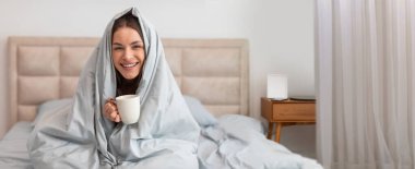 Yumuşak bir battaniyeye sarılı bir kadın, elinde bir kupa kahve tutarken gülümseyerek rahatça yatağa oturur. Doğal ışık odayı doldurur, sıcak ve davetkar bir atmosfer yaratır..