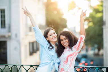 Kutlama için kollarını kaldıran iki arkadaş, gün ışığı bir şehir ortamında ağaçların arasından süzülürken ışıl ışıl gülümsüyor. Kalabalık bir caddenin arka planına mutluluk saçıyorlar..