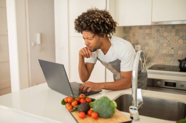 Kıvırcık saçlı siyah bir adam laptopuna odaklanırken şık bir mutfakta taze sebzelerle çevrilidir. Yemek yapmayı ve çalışmayı dengeliyor, modern yaşam tarzı seçimlerini sergiliyor..