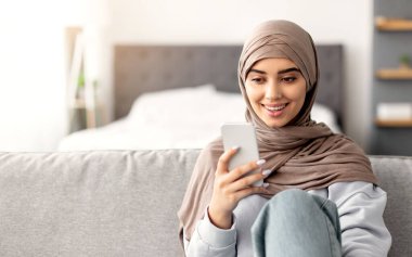 Mobil İletişim. Türbanlı Gülümseyen Müslüman Kadın 'ın Portresi, Akıllı Telefon Kullanımı, Arkadaşlarla Mesajlaşan Olumlu İslamcı Kadın veya Kanepede Oturan İnternet İzleme, Boş Alan