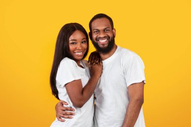 Sevgi dolu bir aile. Güzel, beyaz tişörtlü, sarı turuncu stüdyo arka planında izole bir şekilde duran ve poz veren güzel kadınını kucaklayan yakışıklı siyahi adamın portresi.