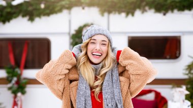 Sıcak bir ceket ve şapka ile sıcak giyinmiş neşeli bir kadın, şenlikli dekorasyonlarla süslenmiş bir kış ortamında parlak bir şekilde gülümsüyor. Karla kaplı çevre neşeli atmosferi güçlendirir..