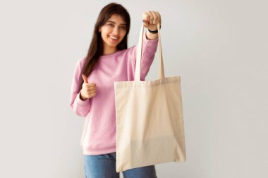 Plastik kullanmayı bırak. Neşeli Milenyum Kadını Kameraya Beyaz Çevre Torbası Gösterip İşaret İşareti Yapıyor. Stüdyo Arkaplan Duvarında Duran Bayan Flax Alışveriş Çantası, Seçici Odaklanma