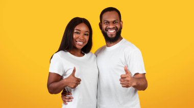 En iyi seçenek, bunu seviyoruz. Afro-Amerikalı bir adamın ve bayanın sarılıp işaret işareti göstermesinin portresi. Millennial ailesi iki kişilik fotoğraf makinesine bakıyor, sarı stüdyo duvarı.