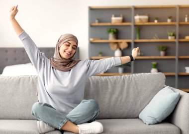 Eğlence için. Oturma odasındaki rahat koltukta oturan güzel İslamcı kadın, kollarını uzatıyor. Başörtüsü takan mutlu bayan, gözleri kapalı dinleniyor, boş zamanın tadını çıkarıyor, işe ara veriyor.