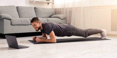 Çevrimiçi antrenman. Genç adam evde, panoramada, boş alanda çevrimiçi öğretmenlerle tahta egzersizi yapıyor.