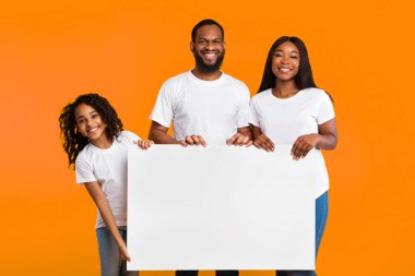 Üç kişilik neşeli siyah aile ve sarı stüdyonun arka planında izole edilmiş kameraya gülümseyen mesajın için boş kağıt afişi gösteriyorlar. Beyaz Meydan Reklamcılık Kurulu
