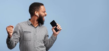 Müzik aşığı. Neşeli zenci erkek modelin portresi şarkı söylüyor ve akıllı telefonunu mavi stüdyo duvarında mikrofon olarak kullanıyor.