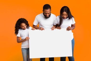 Neşeli Afro-Amerikan erkek, kadın ve kız boş reklam panosuna, sarı stüdyo geçmişine bakıyor. Gülümseyen siyah aile ellerinde reklam için boş tahta ya da metin tutuyor.