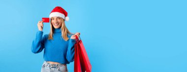İnternetten Noel indirimi. Noel Baba şapkalı mutlu bayan elinde alışveriş çantaları ve kredi kartıyla stüdyoda poz veriyor. Dolu Xmas alışverişi ve banka kartı olan kadın, kopyalama alanı