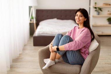 Uyum kavramı. Yatak odasında rahat bir sandalyede oturan, bacaklarını kucaklayan, pencereden dışarı bakan, boşluğu kopyalayan mutlu genç bir kadının portresi. Kadın Boş Zamanın ve Haftasonunun tadını çıkarıyor