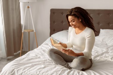 Sıcak bir hafta sonu. Gülümseyen Afrikalı Amerikalı bir kadının portresi rahat bir yatakta oturuyor, kağıt kitap okuyor, boş zamanın tadını çıkarıyor, kopya alanı, seçici odaklanma. Boş zaman ve rahatlama konsepti