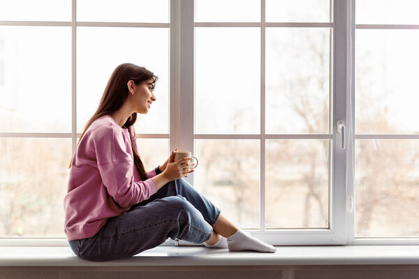 Time For Yourself. Profile side view of calm casual lady sitting on white modern windowsill at home, looking through the glass window, holding cup and drinking hot coffee, thinking. Free copy space
