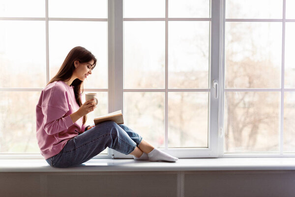 Free Time To Relax. Profile of beautiful woman sitting on white windowsill at home, reading paper book, holding cup and drinking hot coffee. Happy lady taking break with mug near window, copy space