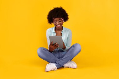 Tablet Bilgisayar kullanarak gülümseyen Afro-Amerikan Kadın Stüdyoda Pembe Arkaplan Üzerinde Oturan İnternet. E-Öğrenme, Ağ ve Çevrimiçi Teknoloji. Gadget Yaşam Tarzı Konsepti