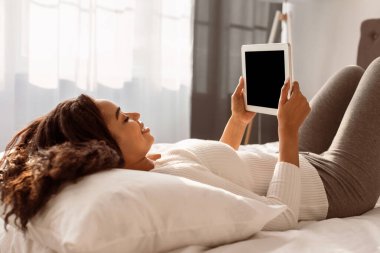 Dinlen, rahatla, Eğlence ve Eğlence Konsepti. Genç Afrikalı Amerikalı Kadın Boş Mokup Ekran Videosu ile Dijital Tablet Kullanıyor, Evde Yastıkta Yatıyor, Hafta sonları Boş Zaman Geçiriyor