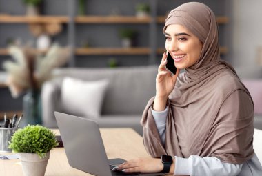 Mobil İletişim. Gülümseyen Müslüman iş kadını cep telefonuyla konuşuyor ve merkez ofiste dizüstü bilgisayarla çalışıyor, bilgisayarını kullanan olumlu bir kadın hoş bir telefon görüşmesi yapıyor, uzak mesafelere gitmekten keyif alıyor.