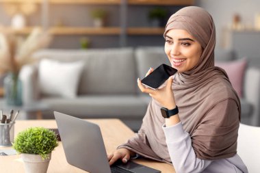 Dijital Hoparlör Uygulaması. Masada oturan, cep telefonuyla konuşan, çevrimiçi arama için dizüstü bilgisayar ve sanal ses asistanı kullanan veya hatırlatıcı kaydeden tesettürlü genç Müslüman kadının portresi