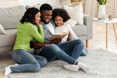 Mutlu Afro-Amerikan ailesi birlikte kitap okuyor, Coronavirus karantinası sırasında evde kalıyor, oturma odasında yerde oturuyor, bağ kuruyor.