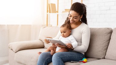 Neşeli Siyah Anne dijital tablet kullanıyor. Küçük bebeği tutuyor. İnternette çizgi filmleri gösteriyor. Oturma odasındaki koltukta oturuyor. Anne Bilgisayar Oyunları Uygulamasında İnternet Taraması Yapıyor