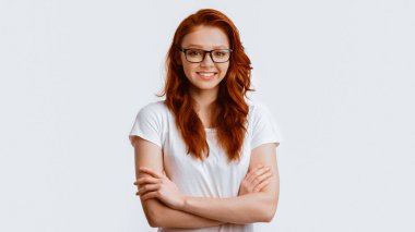 Gözlüklü kızıl saçlı kız stüdyoda kameraya gülümseyen ellerini çaprazlıyor. Panorama