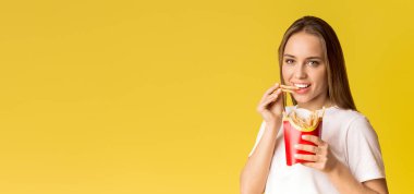 Harmful Food. Portrait Of Happy Young Woman Eating French Fries Potatoes And Looking At Camera, Posing Over Yellow Background With Free Space clipart