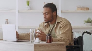 Tekerlekli sandalyedeki genç bir adam ilaçlarını yönetirken dizüstü bilgisayarına odaklanıyor. Bitkiler ve modern dekorlarla çevrili, iyi ışıklandırılmış bir yaşam alanında bir masada oturuyor..