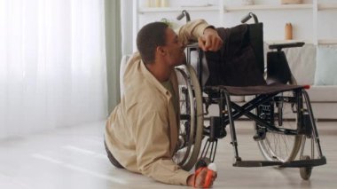 Afrikalı Amerikalı adam evinin zemininde uzanıyor, tekerlekli sandalyesine uzanıyor. Tekerlekli sandalye arkasında sağda..