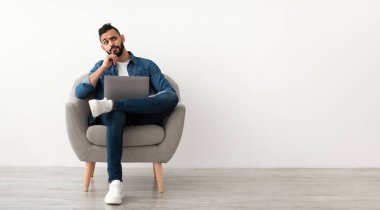 Laptop kullanan, koltukta oturan, beyaz stüdyo duvarına karşı boş alana bakan düşünceli genç bir Arap. Doğu 'lu genç bir adam internette çalışıyor ya da okuyor, bir şeyler düşünüyor.