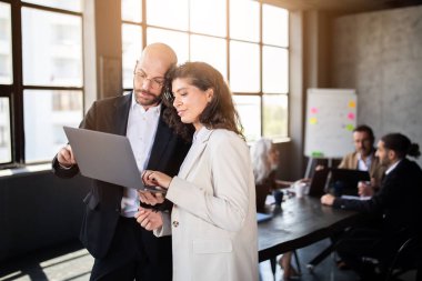 Geniş pencereli çağdaş bir ofiste, iki profesyonel iş arkadaşları arka planda çalışırken bir dizüstü bilgisayarda bilgi tartışıyor. Atmosfer dinamik ve işbirlikçi..