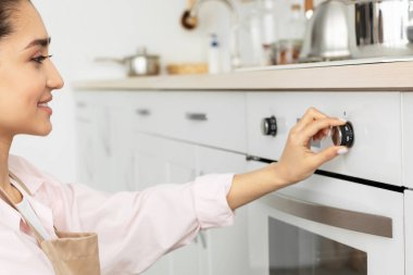 Ev kavramı. Yeni dairesinde, modern mutfakta sobayı kullanan önlüklü, bin yıllık neşeli bir kadının yakın plan portresi. Tutma ve döndürme düğmesi, ocağın sıcaklığını kontrol etme.