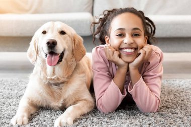 Çocuklar ve Evcil Hayvan. Neşeli zenci kız portresi köpeğiyle yerde yatar, kameraya bakar ve poz verir, oturma odasında ya da yatak odasında altın emeklisiyle dinlenir.