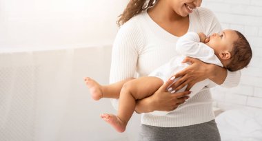 Neşeli siyahi anne kucağında bebeği kucağında tutuyor. Küçük şirin çocuğa gülümsüyor. Evde yatak odasında duruyor. Annelik, annelik izni ve çocuk bakımı kavramı